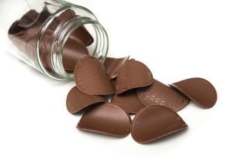 Closeup of chocolate in shaped tiles falling from a glass container on white background