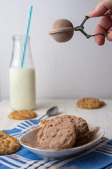 Close-up of woman's hand with chocolate powder falling on plate with chocolate chip cookies, bottle of milk and spoon, on blue napkin, selective focus, vertical