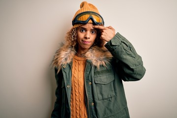 Young african american skier woman with curly hair wearing snow sportswear and ski goggles pointing unhappy to pimple on forehead, ugly infection of blackhead. Acne and skin problem