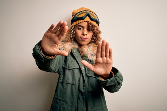 Young african american skier woman with curly hair wearing snow sportswear and ski goggles doing frame using hands palms and fingers, camera perspective