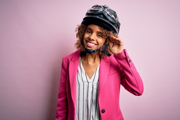 African american motorcyclist woman with curly hair wearing moto helmet over pink background smiling with hand over ear listening an hearing to rumor or gossip. Deafness concept.