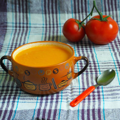 tomato soup in a brown plate with handles, a red spoon and two tomatoes side view on a white and lilac tablecloth .gluten-free vegan food . homemade vegetable soup