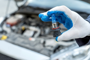 Repairment holding new car light bulb under deception for headlamp, car as background