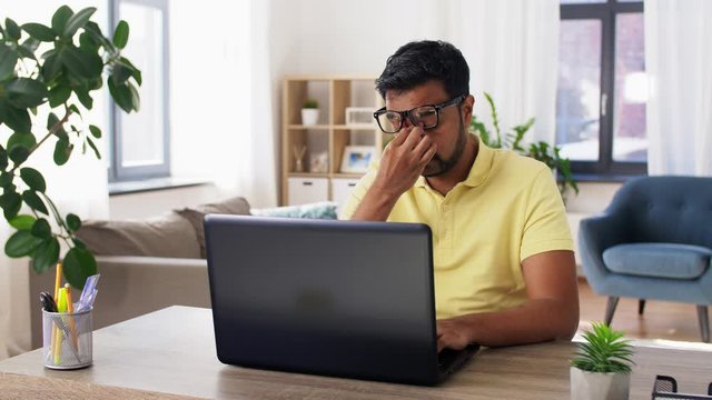 remote job, technology and people concept - tired young indian man in glasses with laptop computer working and rubbing his nose bridge at home office