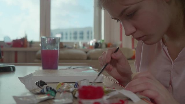 Side view of girl at table near window mixing colours and painting picure.