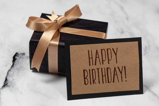 Black gift box with a golden ribbon With a happy birthday Card on a white and black marble table.