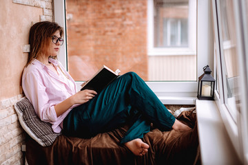 girl in glasses reads a book near the window. Young woman resting on a balcony alone at home