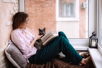 girl in glasses reads a book near the window. Young woman is relaxing on the balcony with her kitten