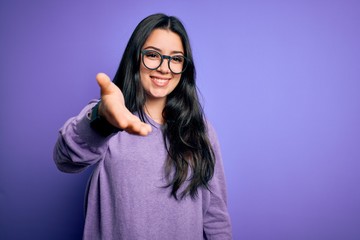 Young brunette woman wearing glasses over purple isolated background smiling friendly offering handshake as greeting and welcoming. Successful business.