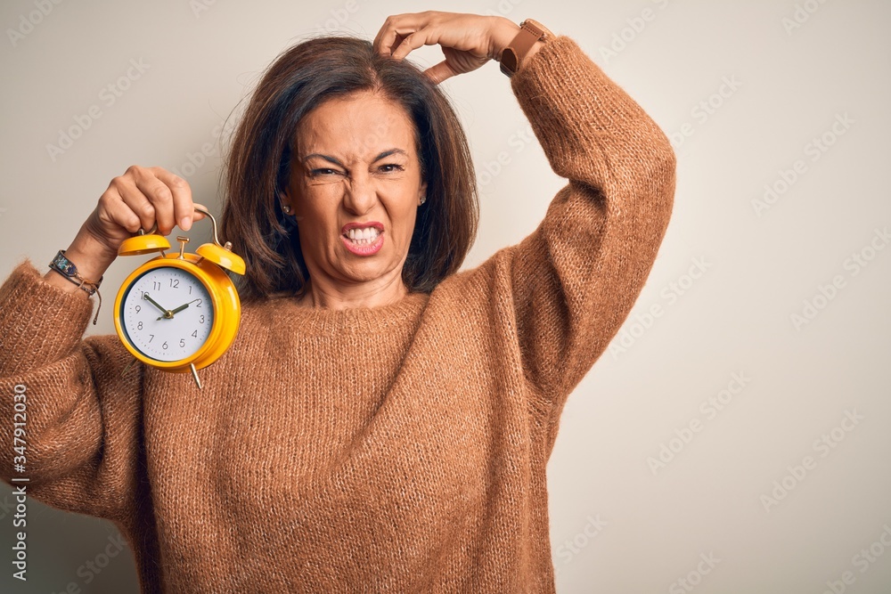 Sticker Middle age brunette woman holding clasic alarm clock over isolated background confuse and wondering about question. Uncertain with doubt, thinking with hand on head. Pensive concept.