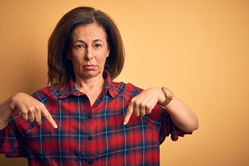 Middle age beautiful woman wearing casual shirt standing over isolated yellow background Pointing down looking sad and upset, indicating direction with fingers, unhappy and depressed.