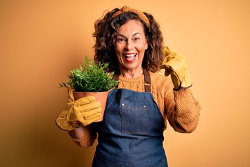 Middle age gardener woman wearing apron holding plant pot over isolated yellow background screaming...