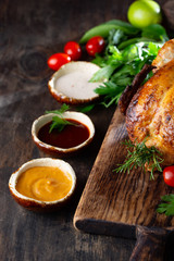 Whole Roasted Chicken on wooden board with rosemary, sauces and vegetables