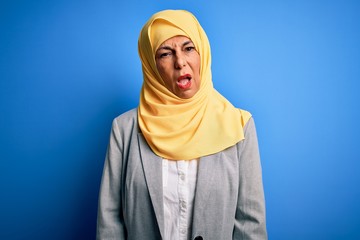 Middle age brunette business woman wearing muslim traditional hijab over blue background In shock face, looking skeptical and sarcastic, surprised with open mouth