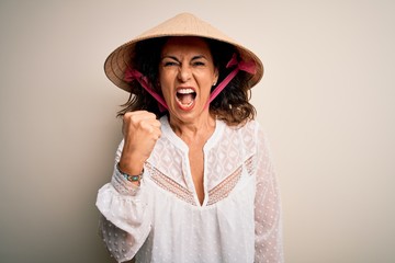 Middle age brunette woman wearing asian traditional conical hat over white background angry and mad raising fist frustrated and furious while shouting with anger. Rage and aggressive concept.