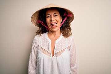 Middle age brunette woman wearing asian traditional conical hat over white background sticking tongue out happy with funny expression. Emotion concept.