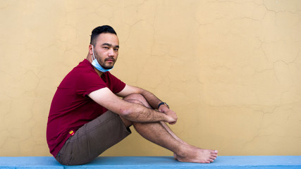 A handsome attractive young Asian man in a face mask during the quarantine, lockdown and self-isolation, sitting on a yellow-brown background, red t-shirt