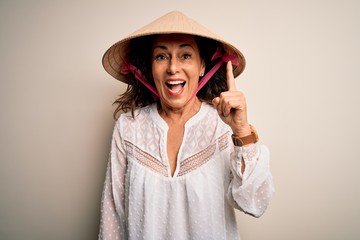 Middle age brunette woman wearing asian traditional conical hat over white background pointing finger up with successful idea. Exited and happy. Number one.