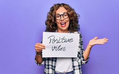 Middle age woman asking for optimistic attitude holding paper with positive vibes message celebrating achievement with happy smile and winner expression with raised hand