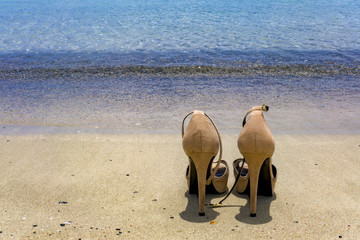Pair of beige high heels shoes left on sandy beach near sea surf. Concept of  travelling, vacation...