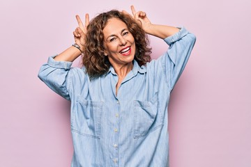 Fototapeta premium Middle age beautiful woman wearing casual denim shirt standing over pink background Posing funny and crazy with fingers on head as bunny ears, smiling cheerful