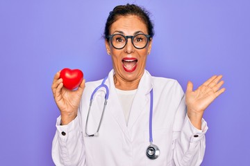 Middle age senior cardiologist doctor woman holding red heart over purple background very happy and excited, winner expression celebrating victory screaming with big smile and raised hands