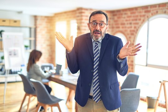Middle age handsome businessman wearing glasses   standing at the office clueless and confused expression with arms and hands raised. Doubt concept.