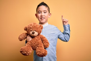 Young little boy kid hugging teddy bear stuffed animal over yellow background surprised with an idea or question pointing finger with happy face, number one