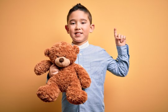 Young little boy kid hugging teddy bear stuffed animal over yellow background surprised with an idea or question pointing finger with happy face, number one