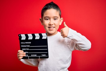 Young little boy kid filming video holding cinema director clapboard over isolated red background...