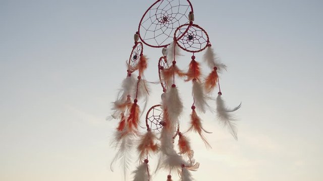 Dreamcatcher with orange feathers, swaying with light wind slow motion against background blue sky at sunset light in in summer. Boho style in spring on sunny day. Indian style amulet. Vintage. Macro