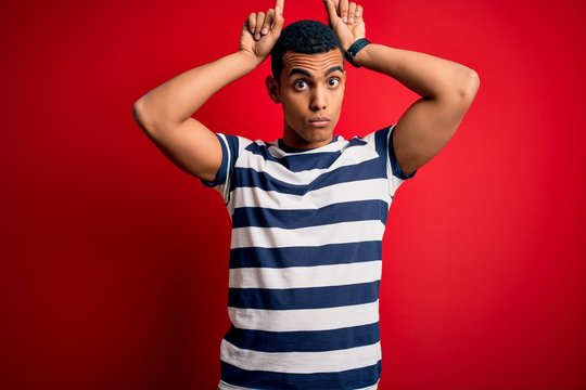 Handsome african american man wearing casual striped t-shirt standing over red background doing funny gesture with finger over head as bull horns