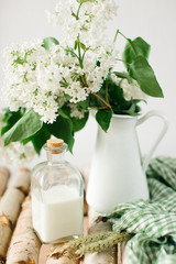 Still life in bright colors, with green accents. Bouquet of white lilac in white vase, next to bottle of milk.