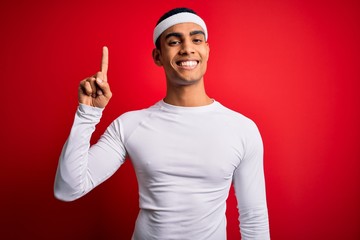 Young handsome african american sportsman wearing sportswear over red background showing and pointing up with finger number one while smiling confident and happy.