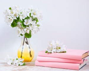Blooming apple tree branches in a glass vase and pink books on a light background. Home interior with spring decor.