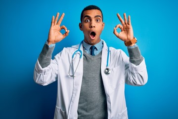 Handsome african american doctor man wearing coat and stethoscope over blue background looking surprised and shocked doing ok approval symbol with fingers. Crazy expression