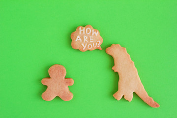 Homemade cookies in shapes of dinosaur and man with inscription ‘How are you?’ on green background, top view. Sweet shortbread with white glaze.