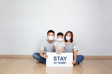 Asian family wearing protective medical mask for prevent virus Covid-19 holding white paper with stay at home on hand and sitting together on floor at home