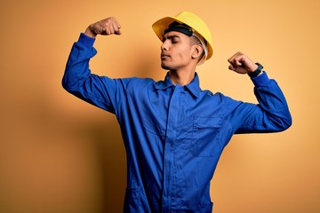 Young handsome african american worker man wearing blue uniform and security helmet showing arms...