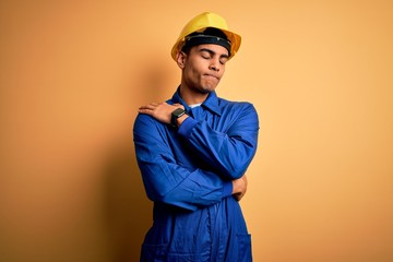Young handsome african american worker man wearing blue uniform and security helmet Hugging oneself...