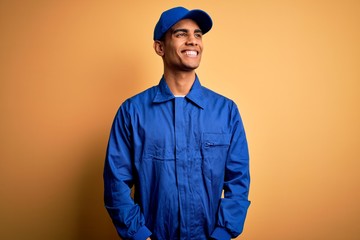 Young african american mechanic man wearing blue uniform and cap over yellow background looking...