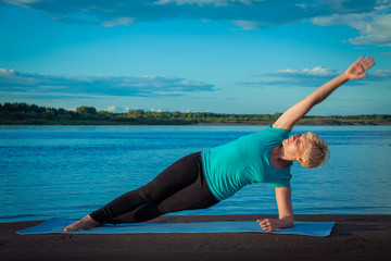 Blond-skinned blonde in a tracksuit on a blue rug on a sandy beach near the river at sunset makes a side plank