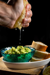 Hand squeezing a lemon to season the delicious and fresh Mexican guacamole sauce, in a blue glass bowl. On a black background