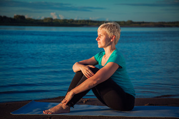 blonde woman with a short haircut on a sandy beach near the water sits on a blue rug and looks into the distance