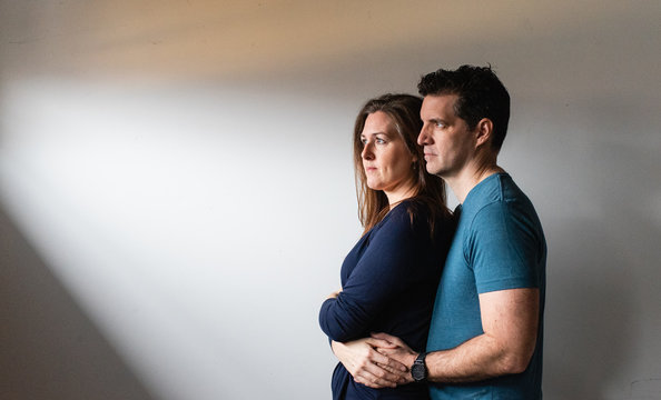 Couple standing close together in a patch of light against white wall.