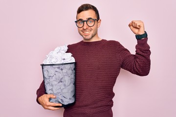 Handsome man with blue eyes wearing glasses holding full paper bin with crumpled papers screaming...