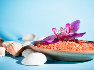 Orange bath salt in a saucer with shells and flower on a blue background with a shadow from a...