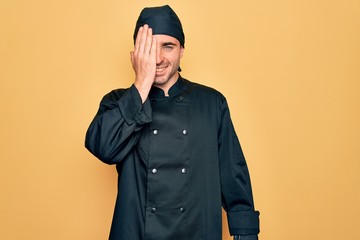 Young handsome cooker man with blue eyes wearing uniform and hat over yellow background covering one eye with hand, confident smile on face and surprise emotion.