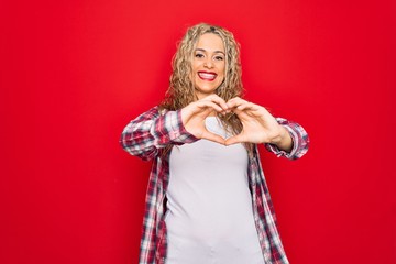 Young beautiful blonde woman wearing casual shirt standing over isolated red background smiling in love doing heart symbol shape with hands. Romantic concept.