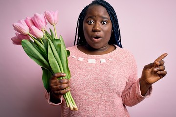 Young african american plus size woman with braids holding bouquet of pink tulips flower Surprised pointing with finger to the side, open mouth amazed expression.